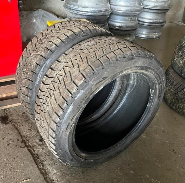 Two black tires stacked on top of each other, with tread visible. Other tires and rims in the background.