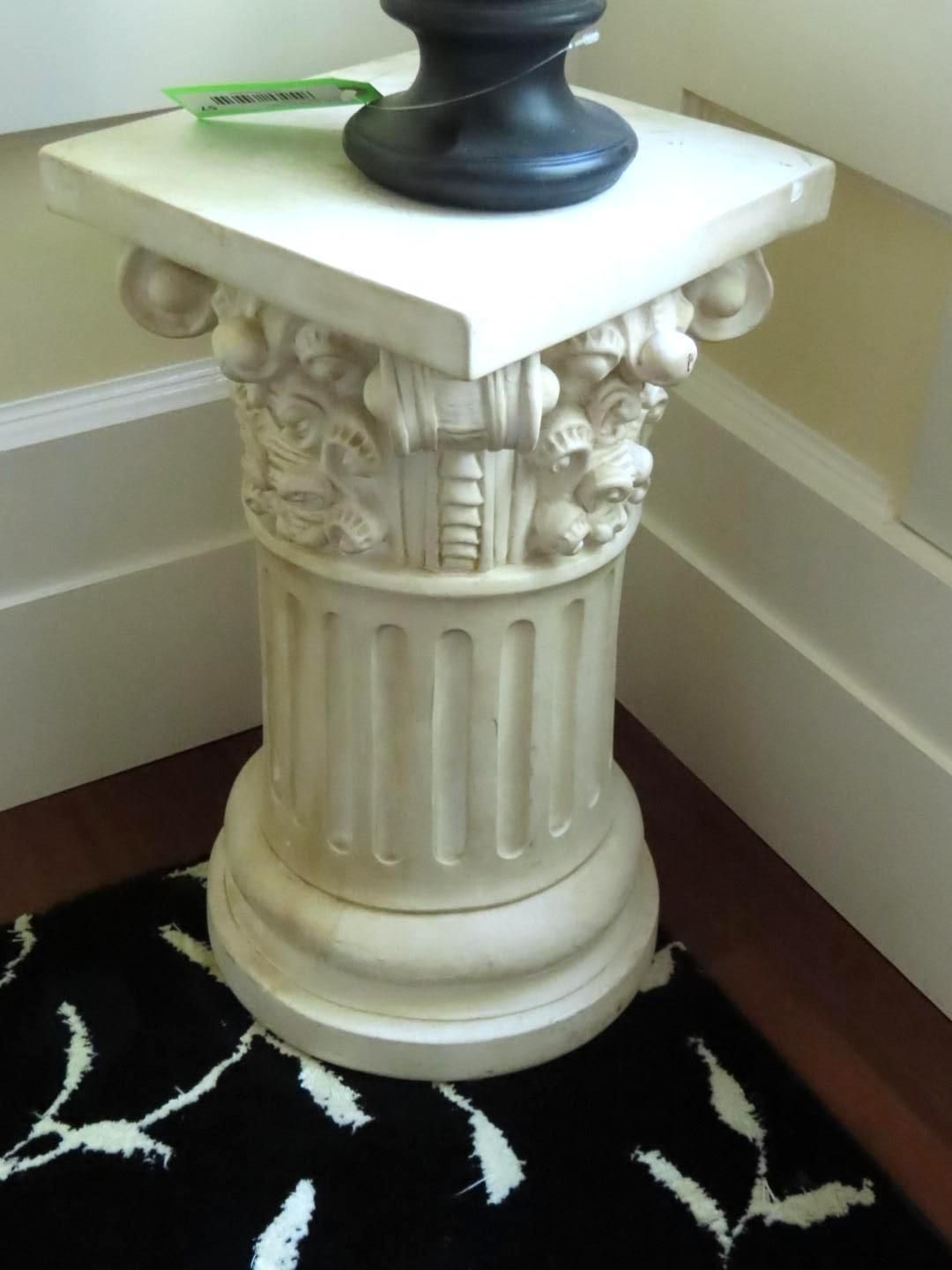 Cream-colored decorative pillar holding a black object, with ornate carving and a square top, on a patterned rug.
