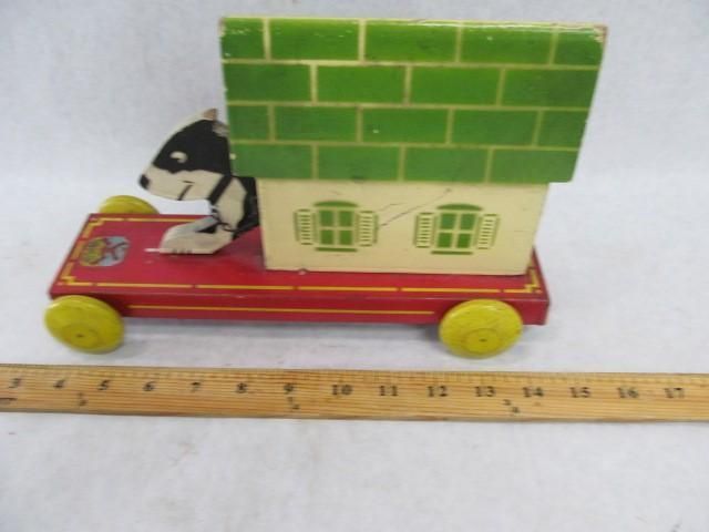 Wooden toy dog driving a red car with a green brick-style roof. Yellow wheels and a ruler are included.