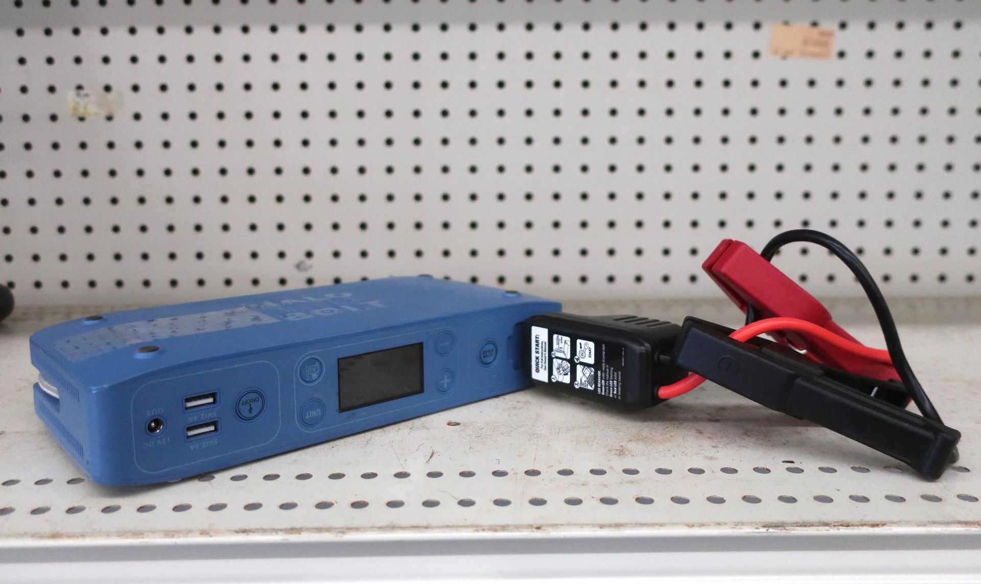 Blue portable car jump starter with attached black clamps on a shelf.