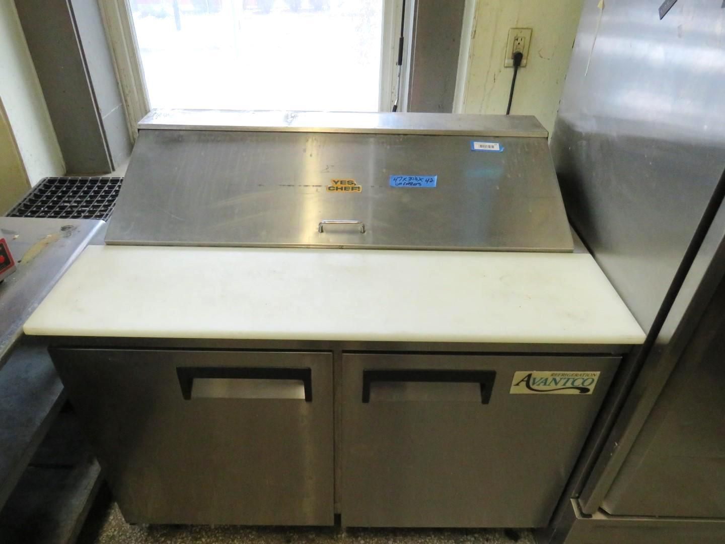 Stainless steel food prep station with cutting board top and refrigerator below, inside a kitchen.