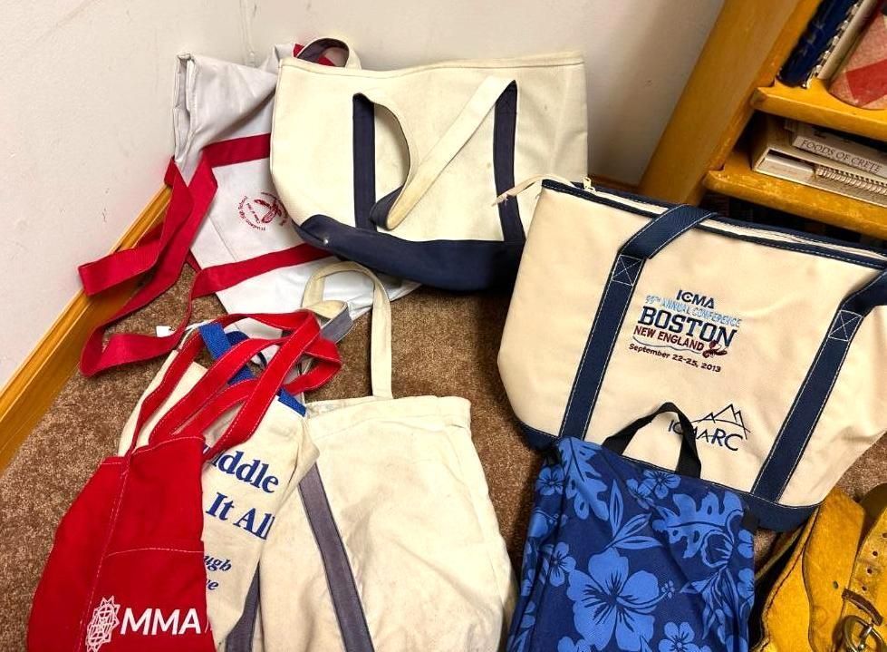 Various tote bags in different colors and styles, some with logos, on a floor near a bookshelf.