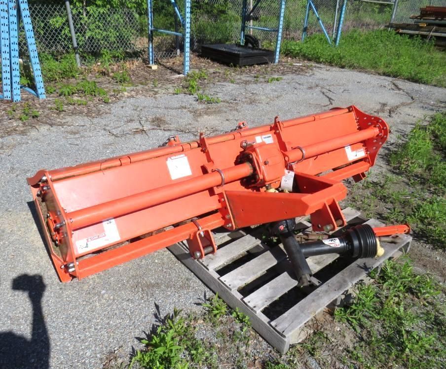 Orange rotary tiller on a wooden pallet, likely for use with a tractor, outdoors on pavement.