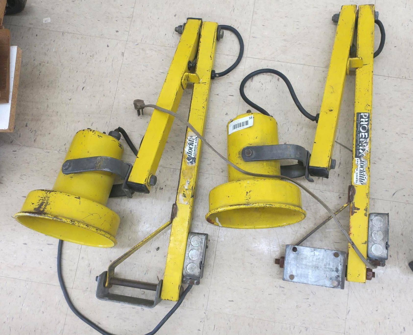 Two yellow industrial clamp lights with cords, lying on a concrete floor alongside metal parts.