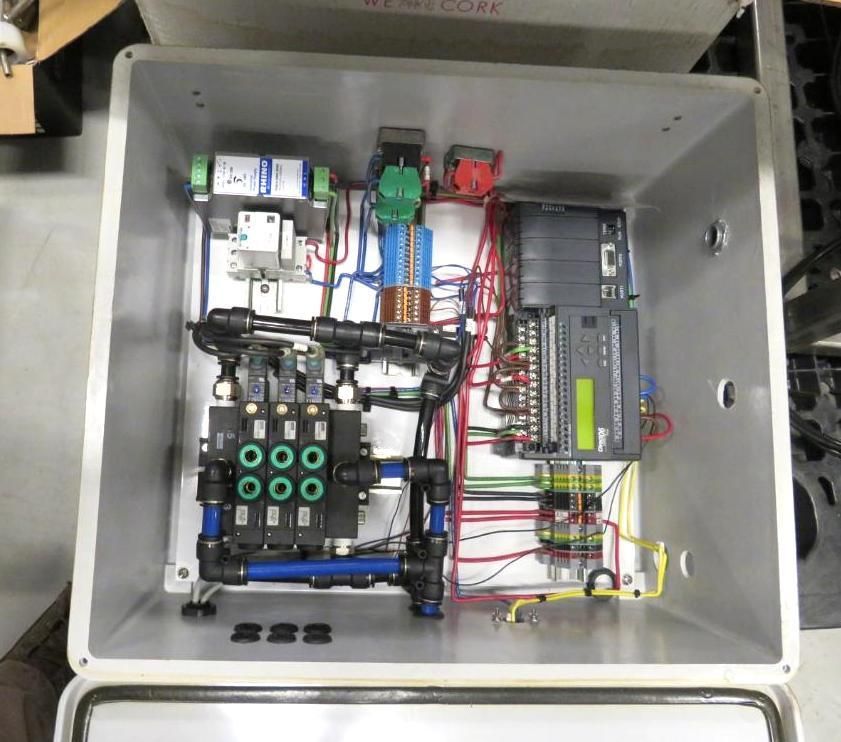 An open metal electrical control box with wired components, including a PLC, valves, and pneumatic tubing.