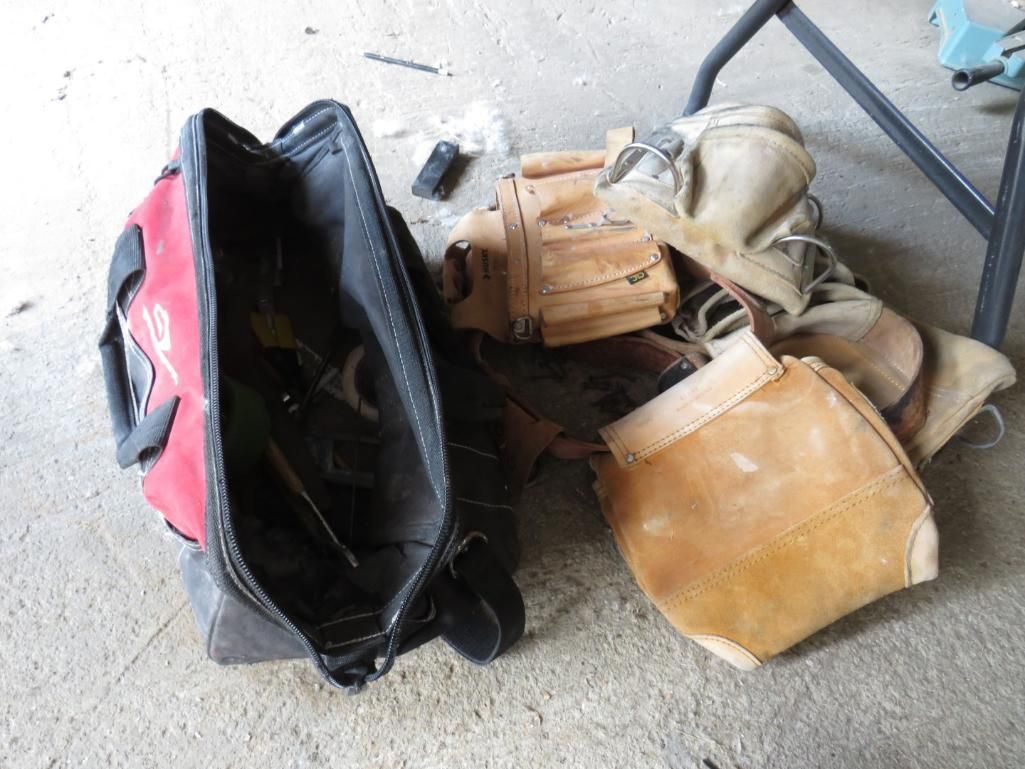 Red and black tool bag with open interior next to a pile of leather tool pouches, on a concrete surface.