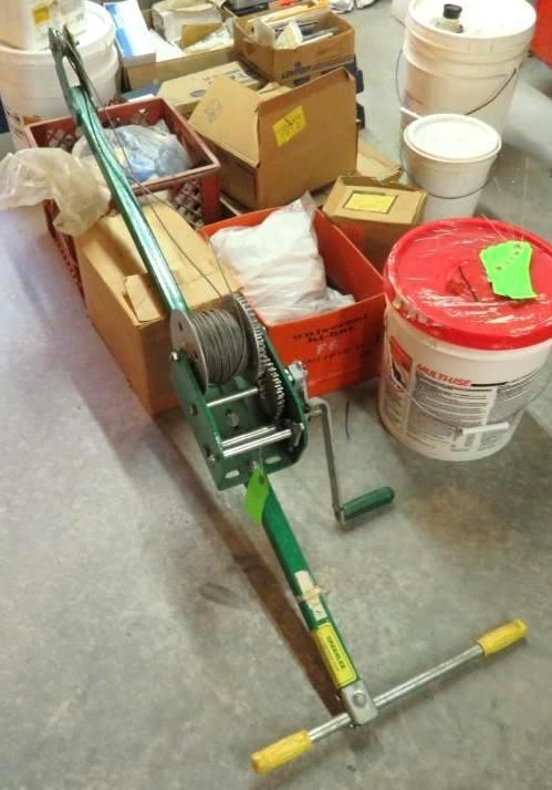 Green hand winch with cable lying on concrete floor surrounded by boxes and buckets.