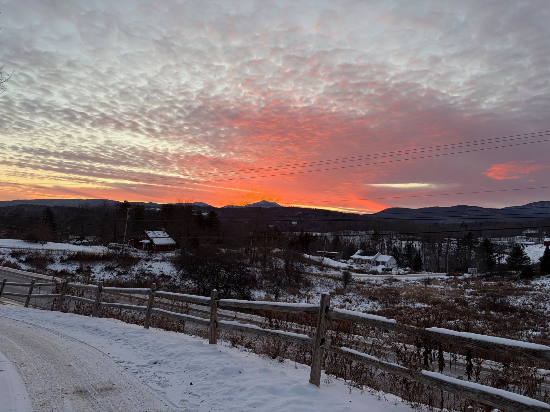 Snowy rural landscape at sunset with a glowing pink-orange sky and wooden fences in the foreground