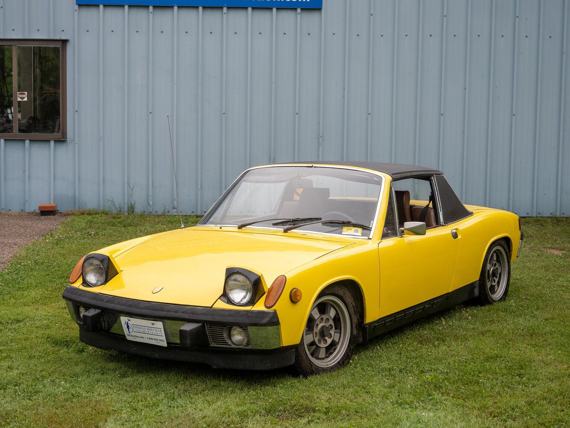 Yellow Porsche 914 convertible parked on grass in front of a blue building with a black top.