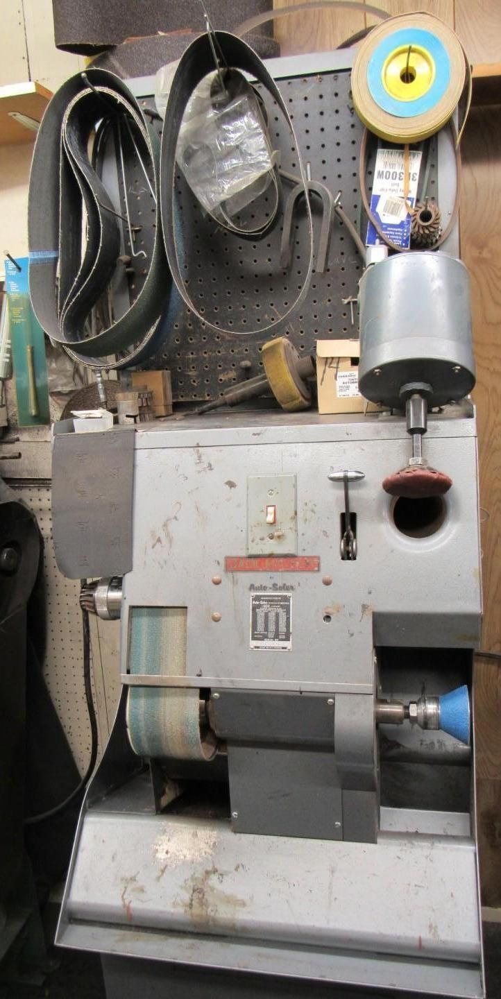 A shoe shining machine in a workshop setting, with belts hanging above, and various polishing heads.