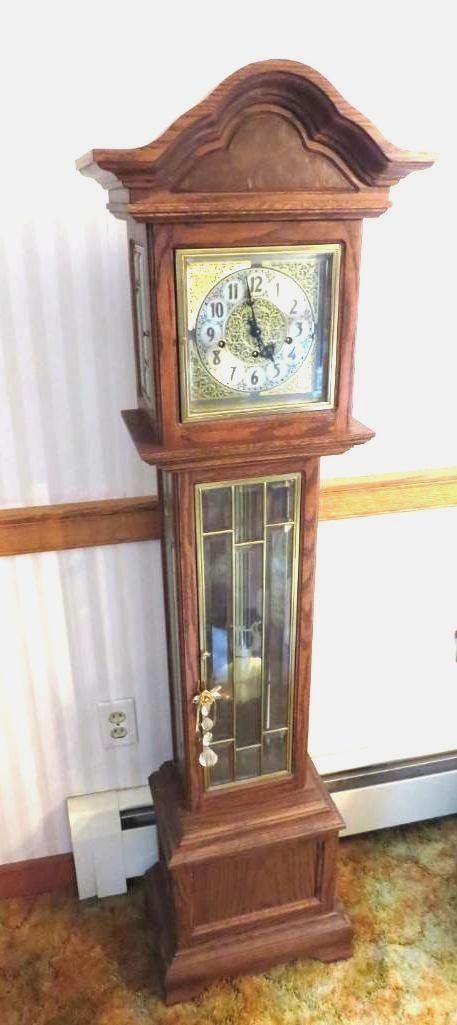 Wooden grandfather clock. It has a square clock face, glass-doored body, and stands on a base.