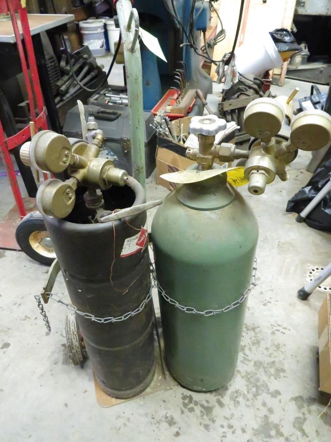 Two gas cylinders with gauges and chains, in a workshop. One is green, the other black.