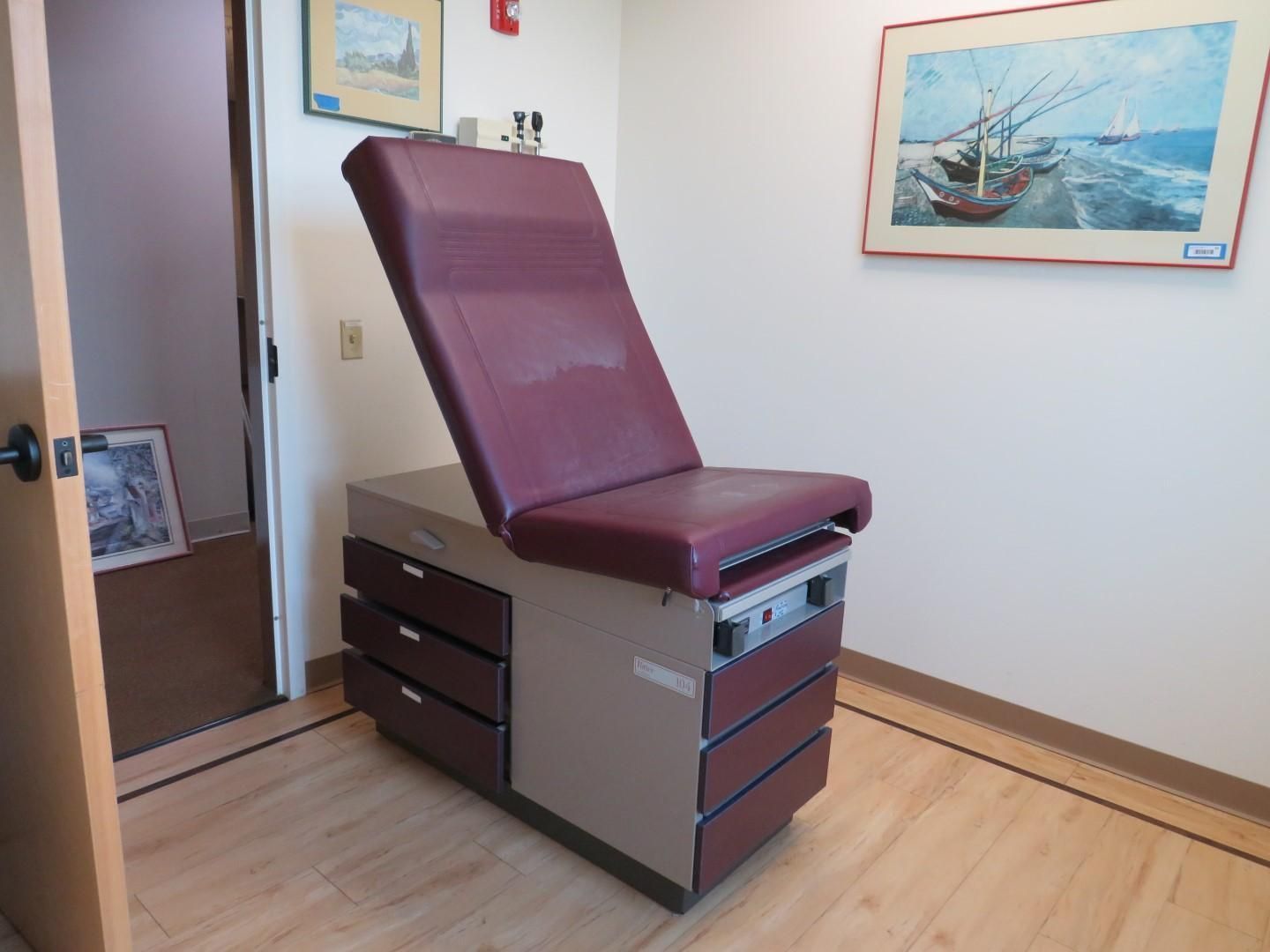 Maroon upholstered medical exam table, drawers, and art on the walls.