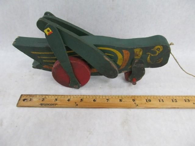 Green wooden grasshopper pull toy with red wheels and yellow accents, beside a wooden ruler.
