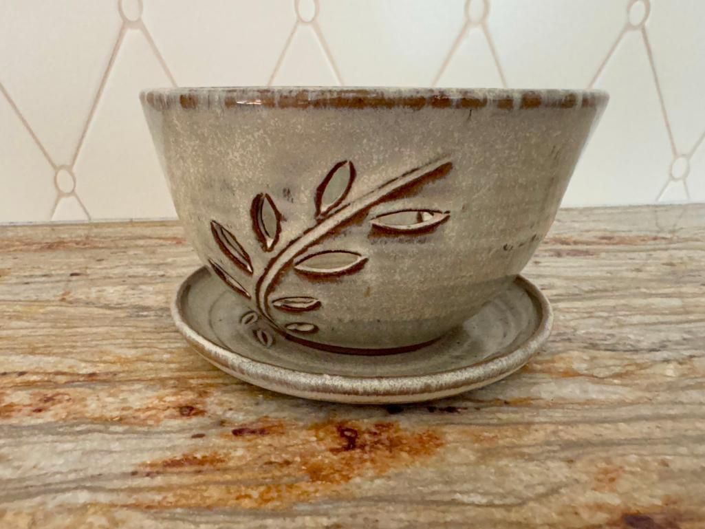 Ceramic bowl planter with leaf design, set on a saucer. Beige-toned, on a patterned surface.