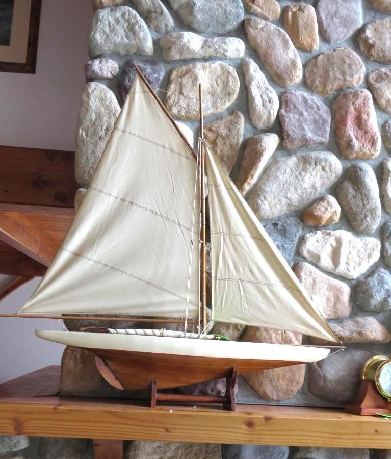 Model sailboat with cream-colored sails and wooden hull, displayed on a wooden shelf in front of a stone wall.