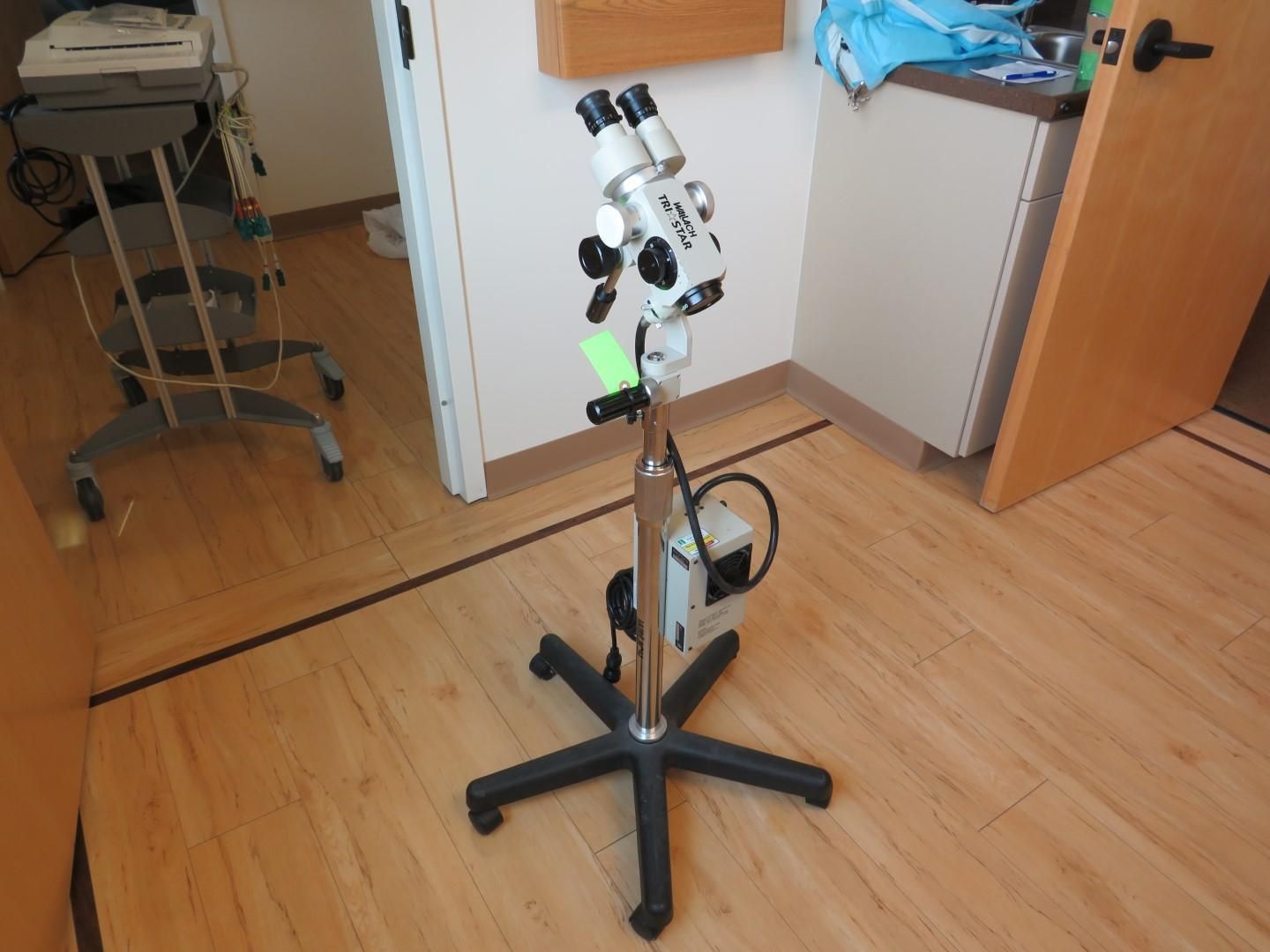 Medical colposcope on a stand in a room with a wooden floor and open door.