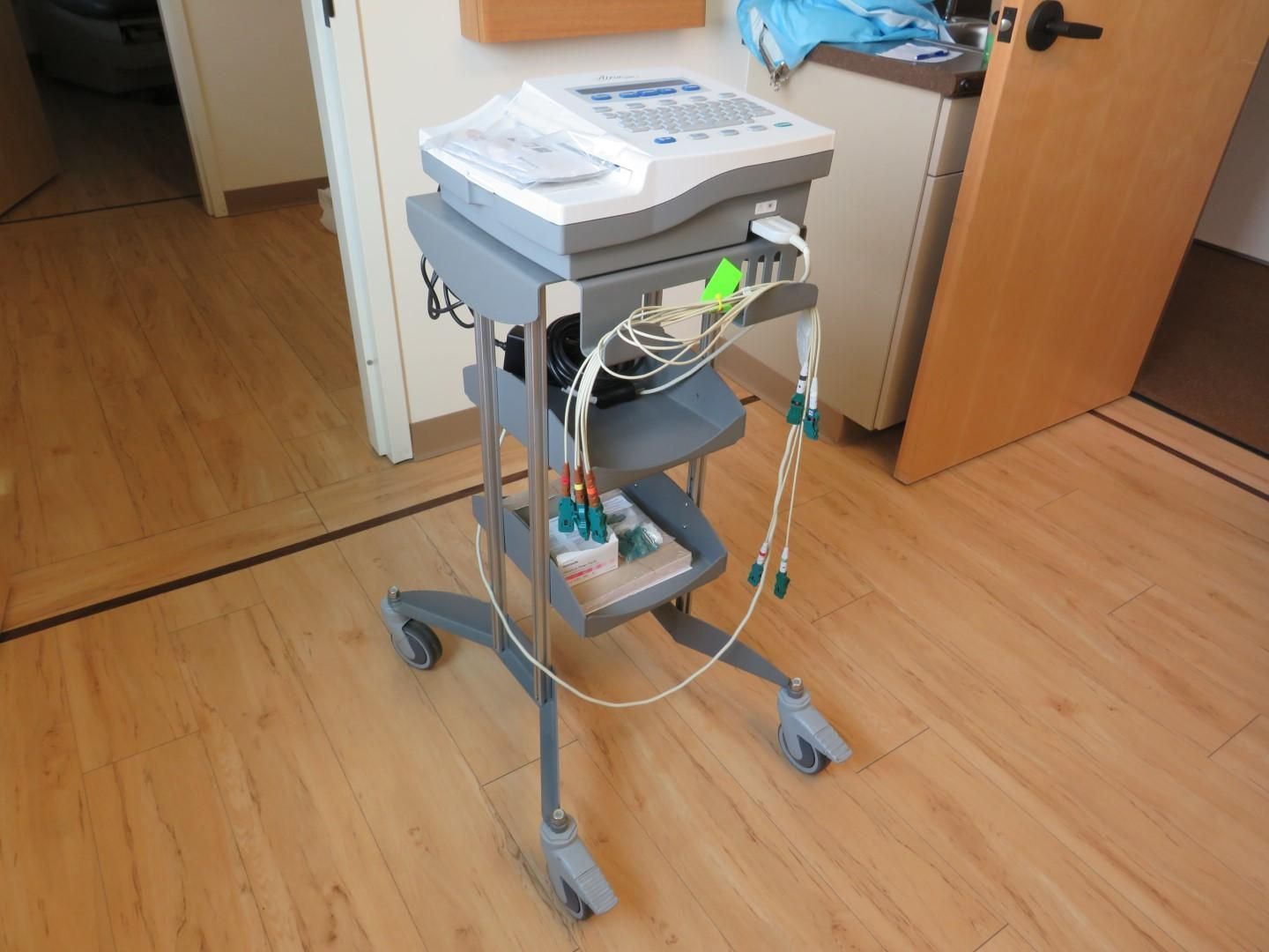 Medical device on a rolling cart in a room with hardwood floors. The device has wires attached.