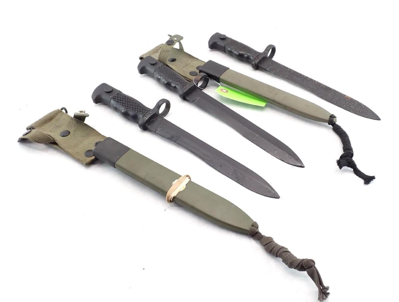 Five bayonet knives in green sheaths and black handles, laid on white background.