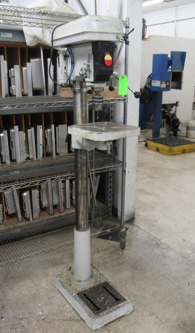 Gray drill press machine in a workshop, next to a shelf with metal items.