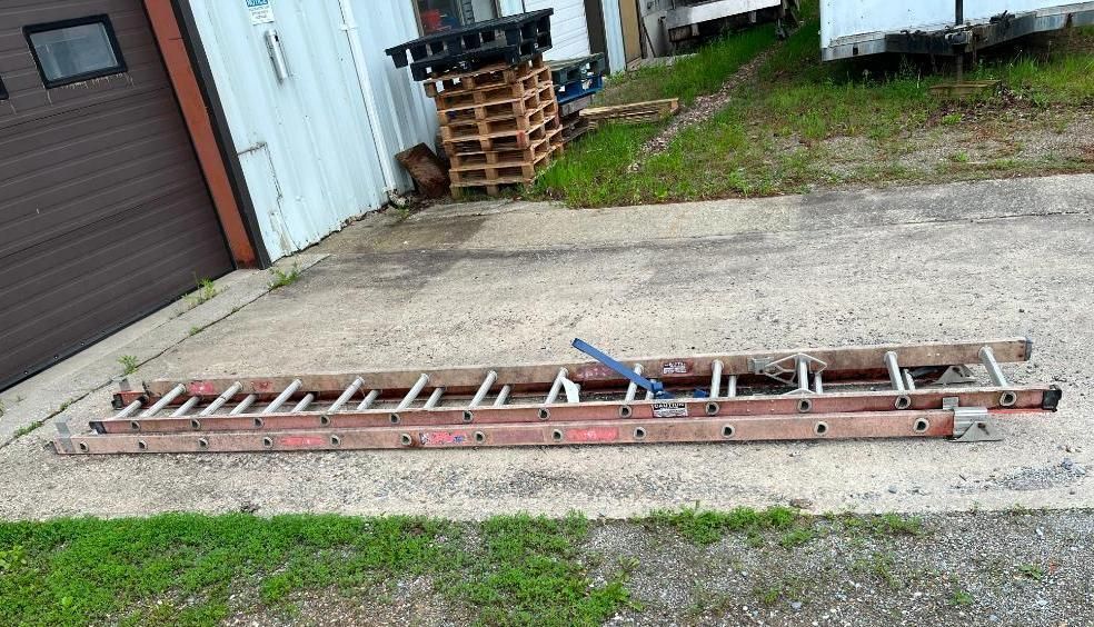 Long red ladder lying on a concrete surface next to a building and grass.