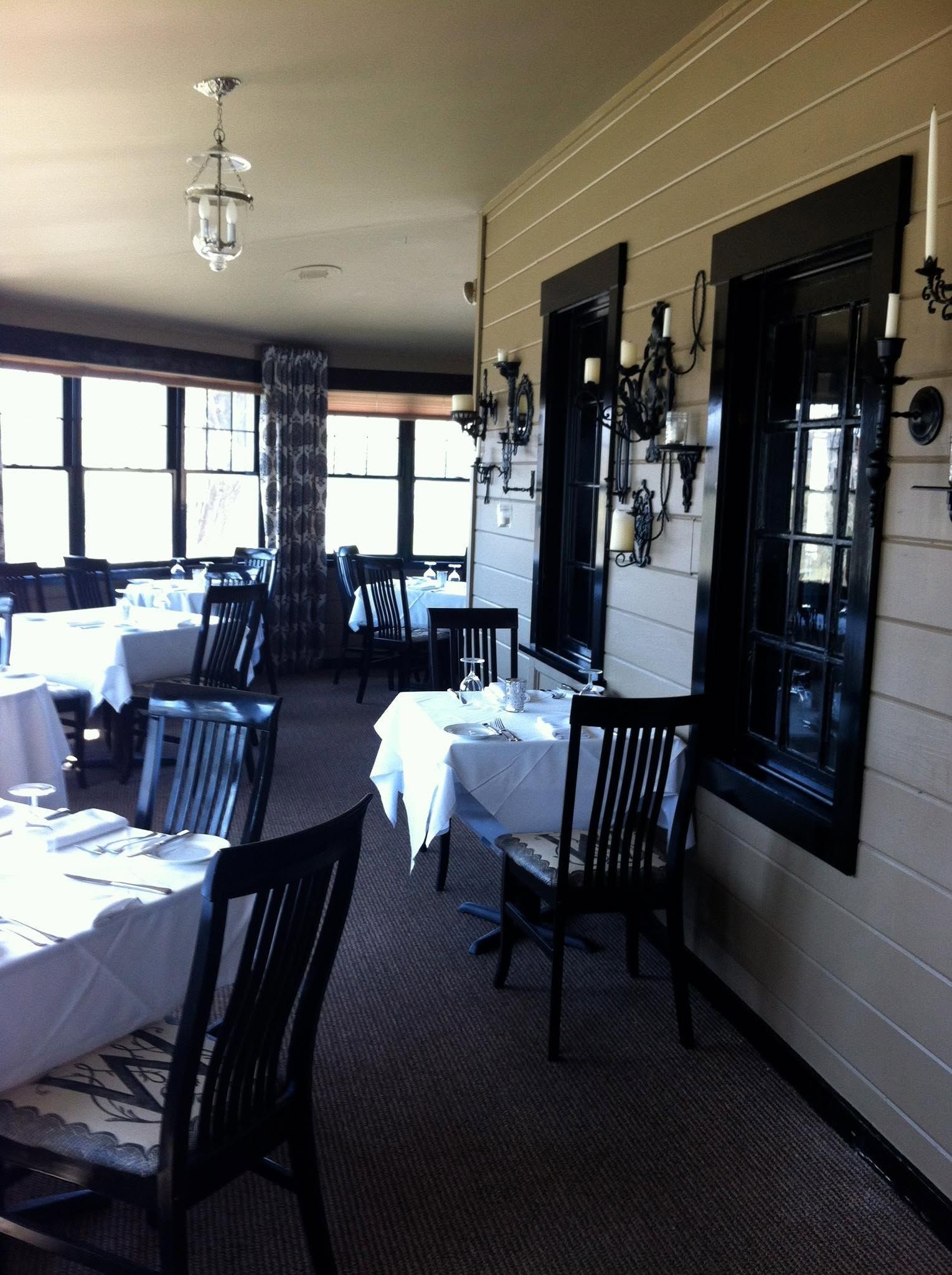 Restaurant dining room with white tablecloth tables, dark chairs, and windows along the wall