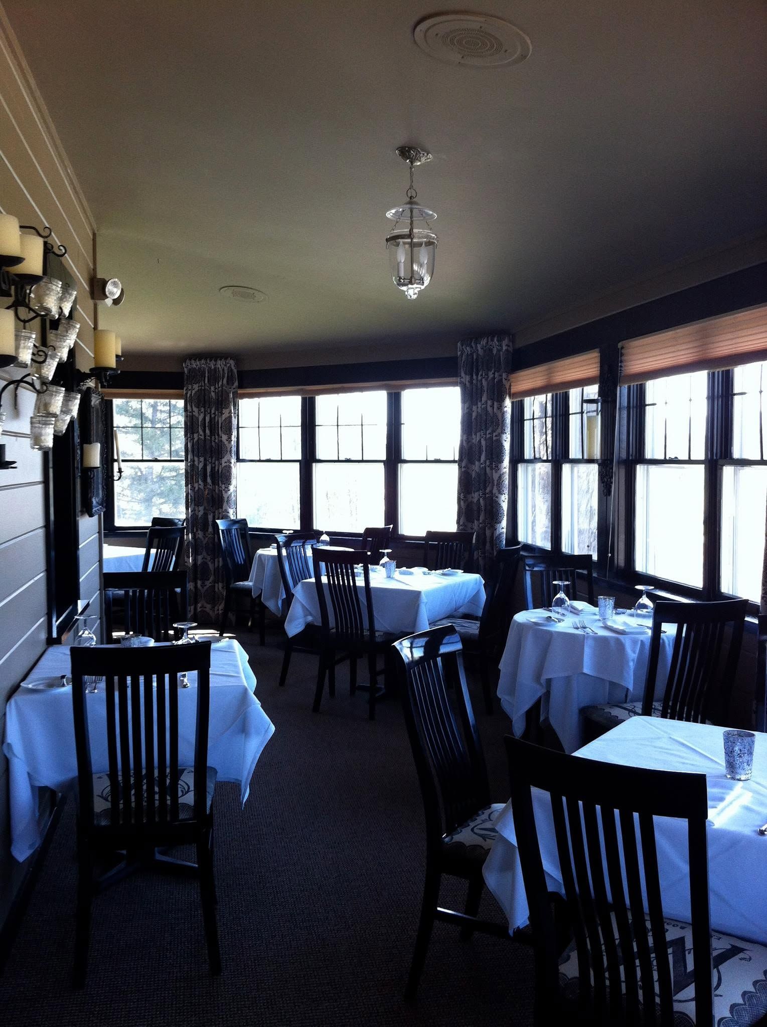 Empty dining room with white tablecloths and black chairs by large windows.