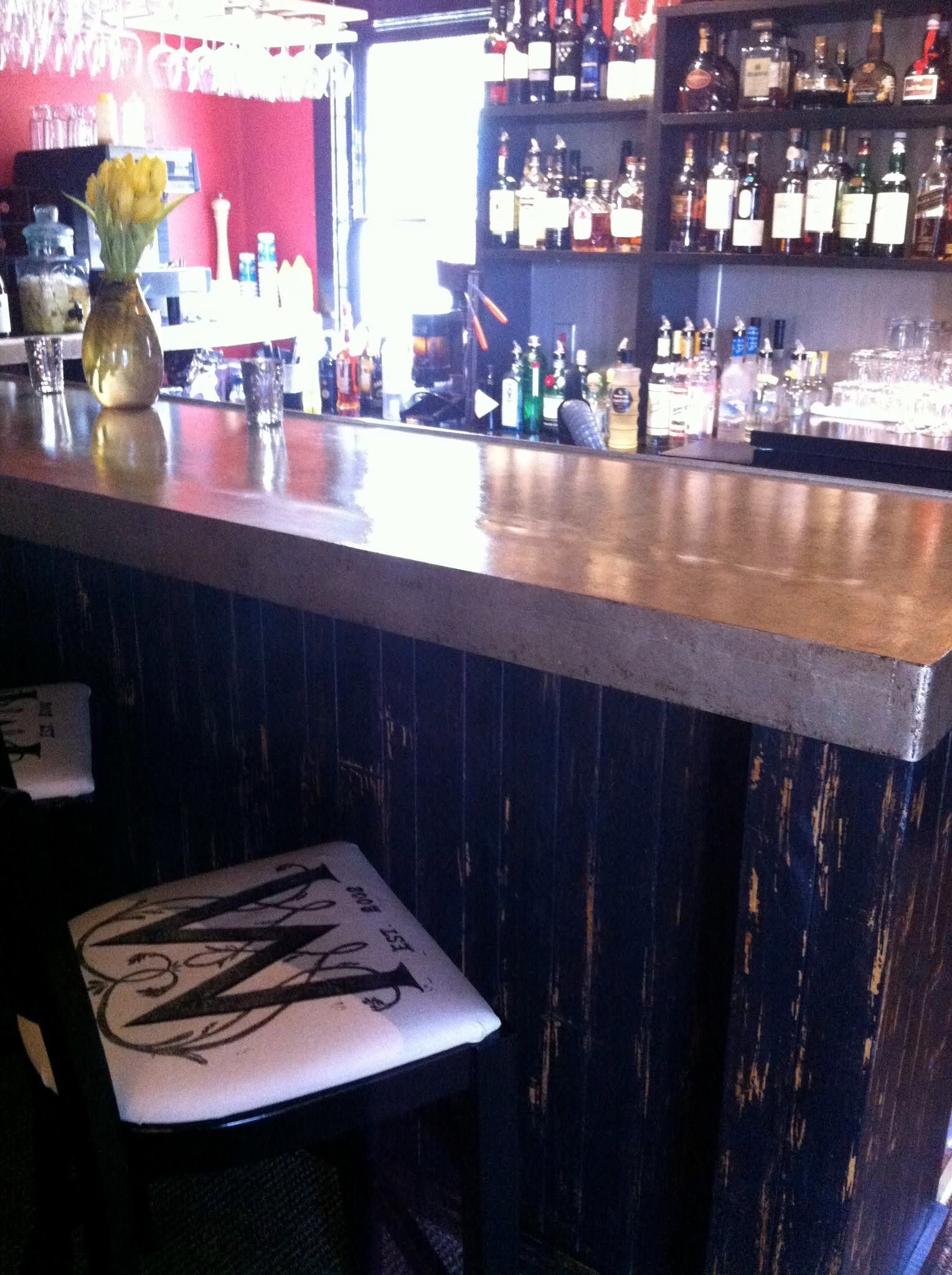 bar interior with a wooden counter, liquor bottles on shelves, and a white stool below.