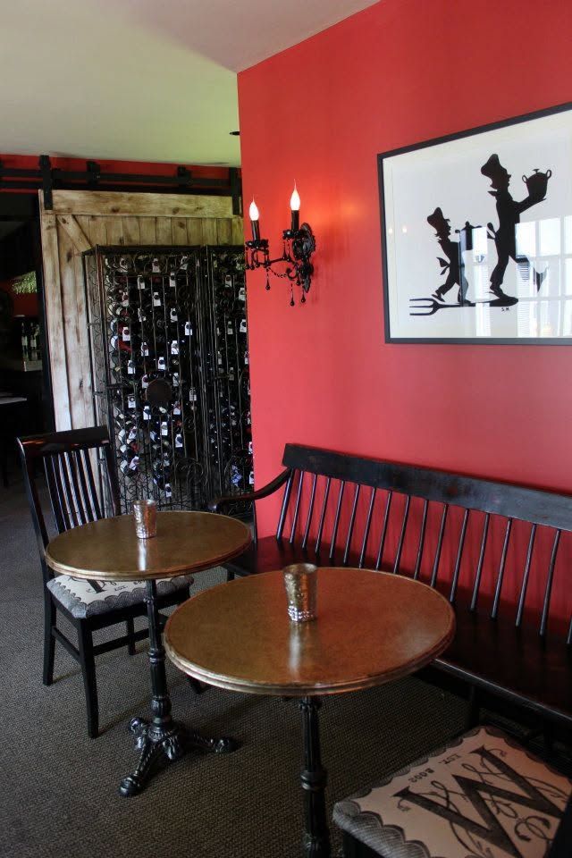 Small cafe corner with two wooden tables, black chairs, red wall, and framed art.