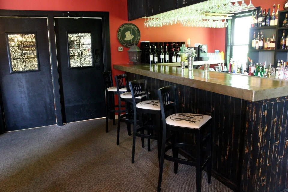 Empty bar interior with black stools, wooden counter, red walls, and glass shelves behind the bar
