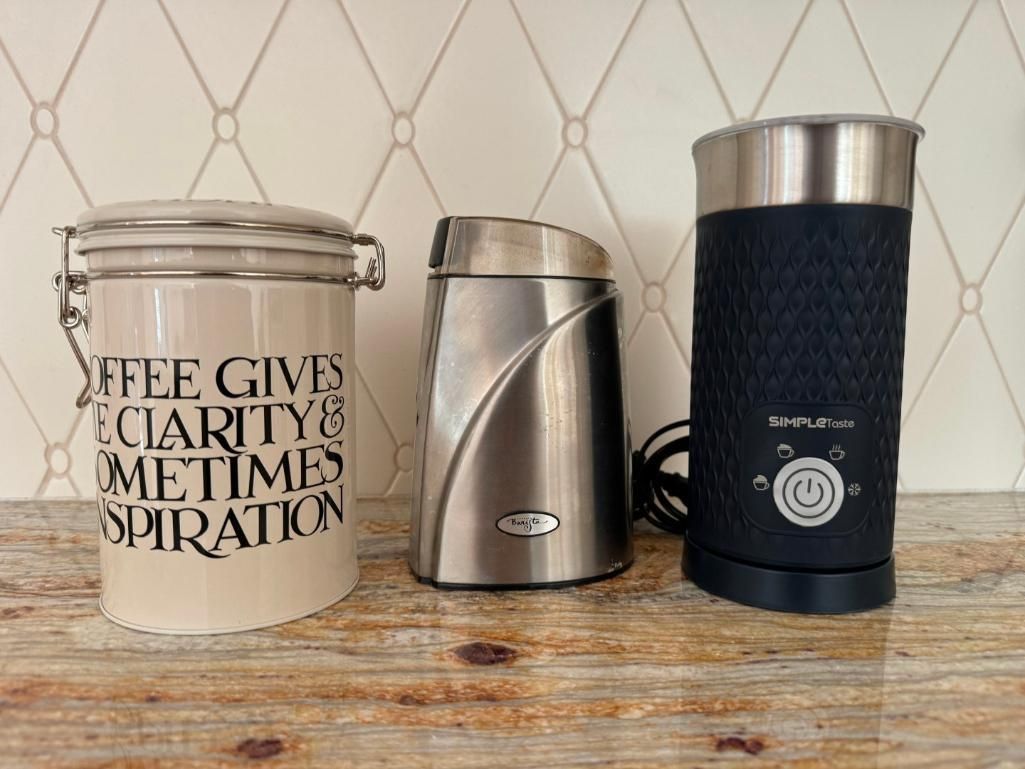 A coffee canister, grinder, and milk frother sit on a granite countertop in front of a tile backsplash.