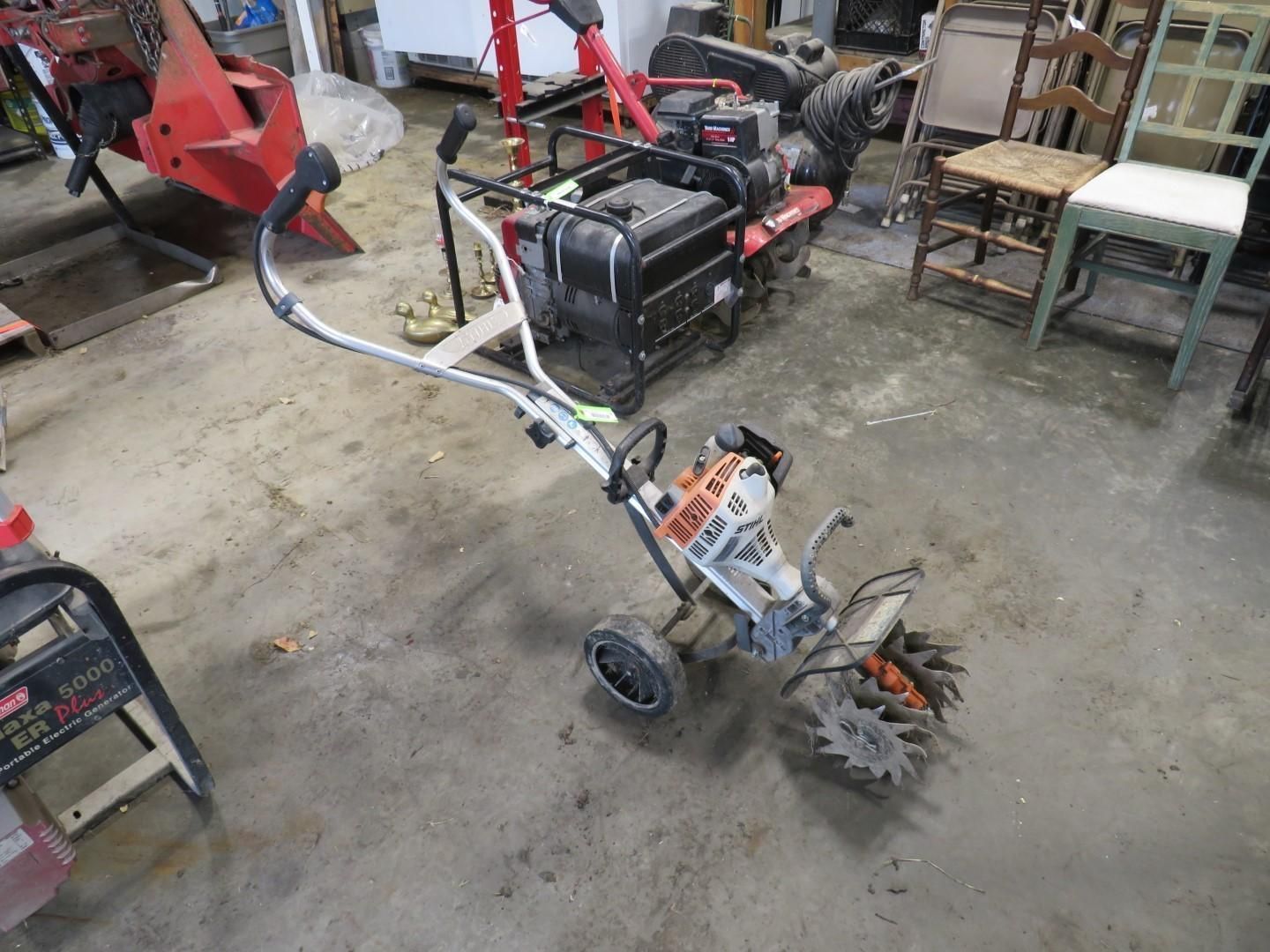 A small garden tiller with a black engine and silver handlebars sits on a concrete floor.