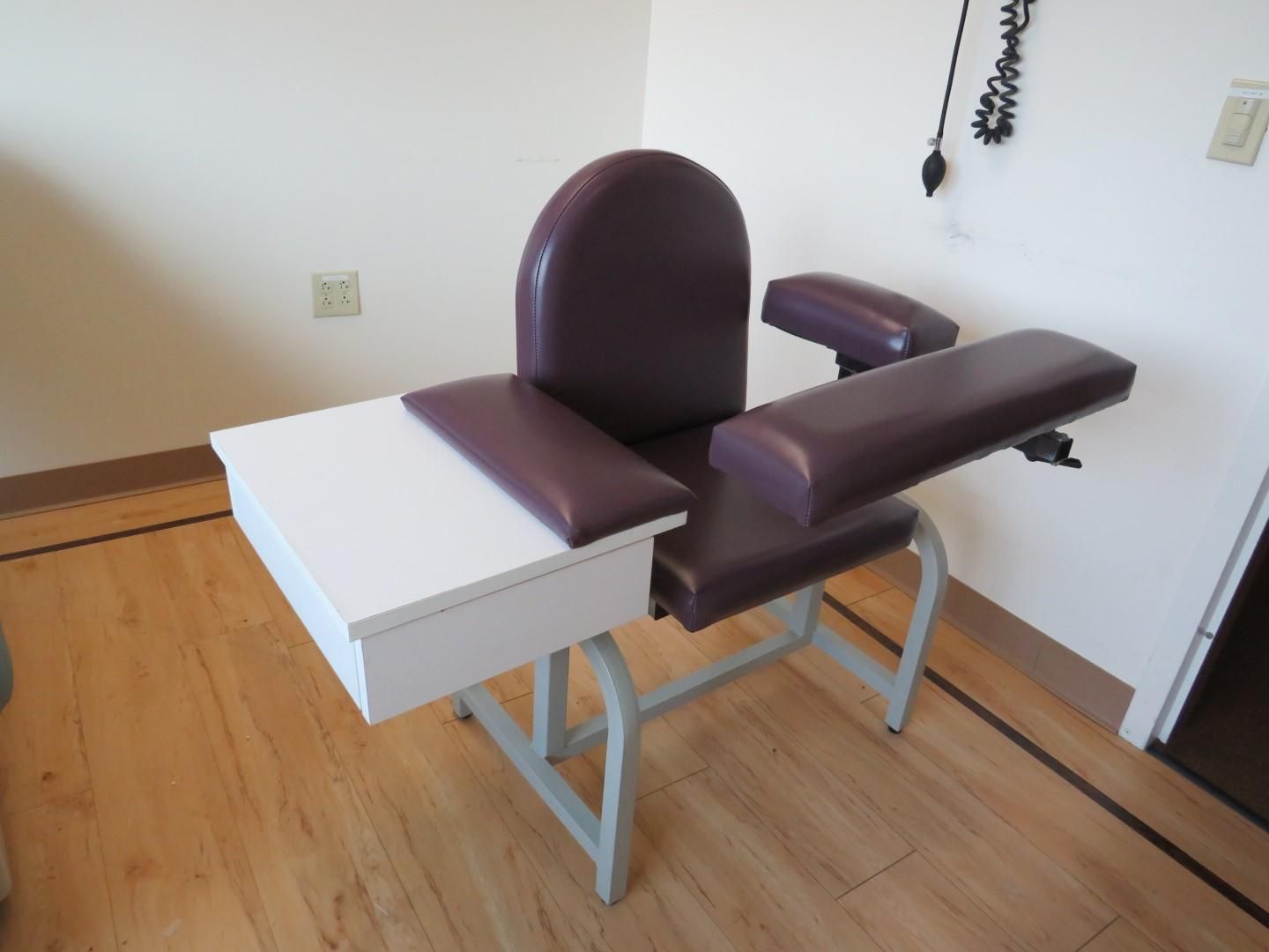 Blood donation chair with armrests, attached tray, and purple upholstery, in a room with hardwood floors and a wall outlet.