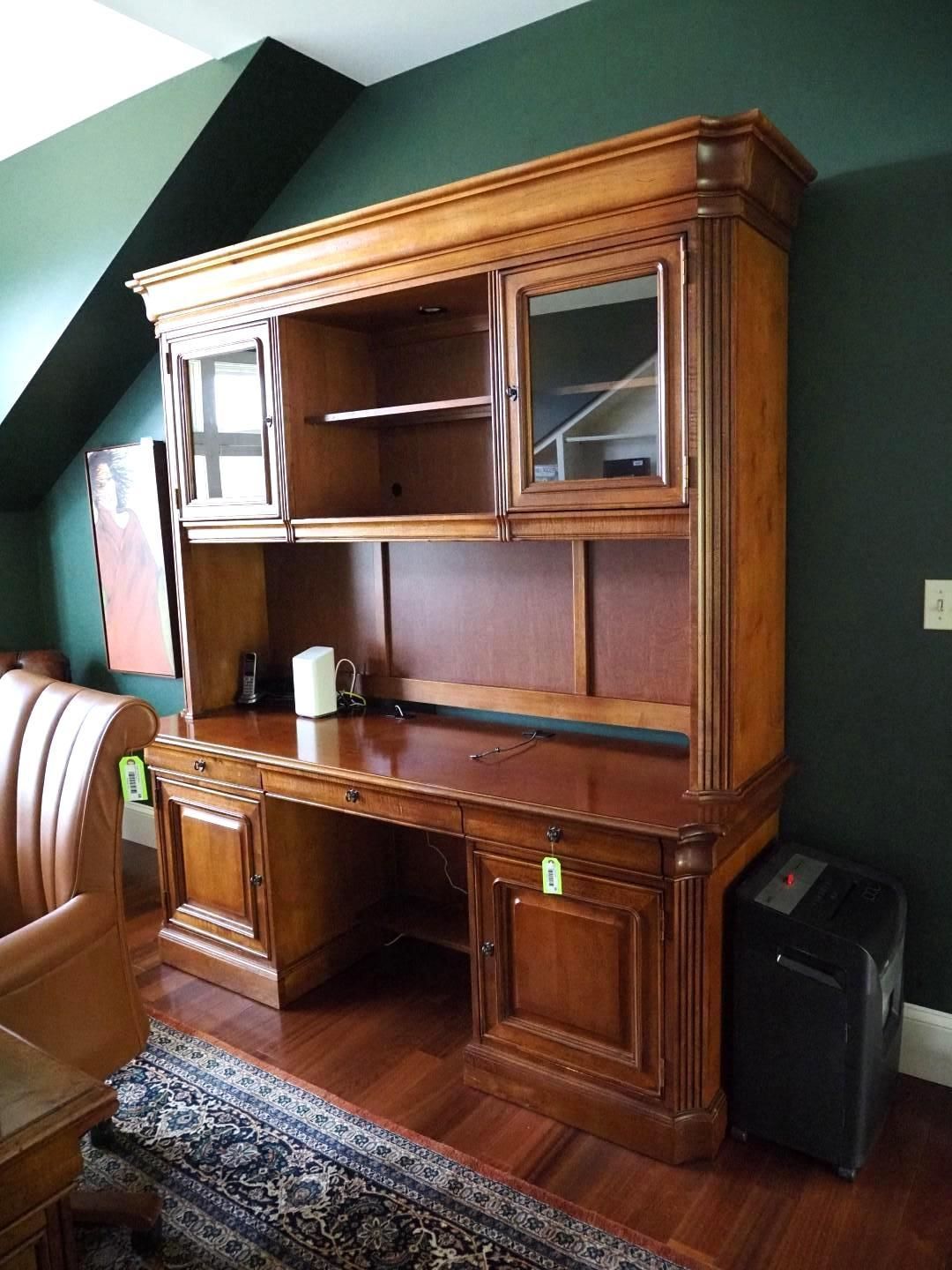 Wooden desk with hutch. Dark wood, two glass-fronted cabinets. Set against a dark green wall, on a patterned rug.