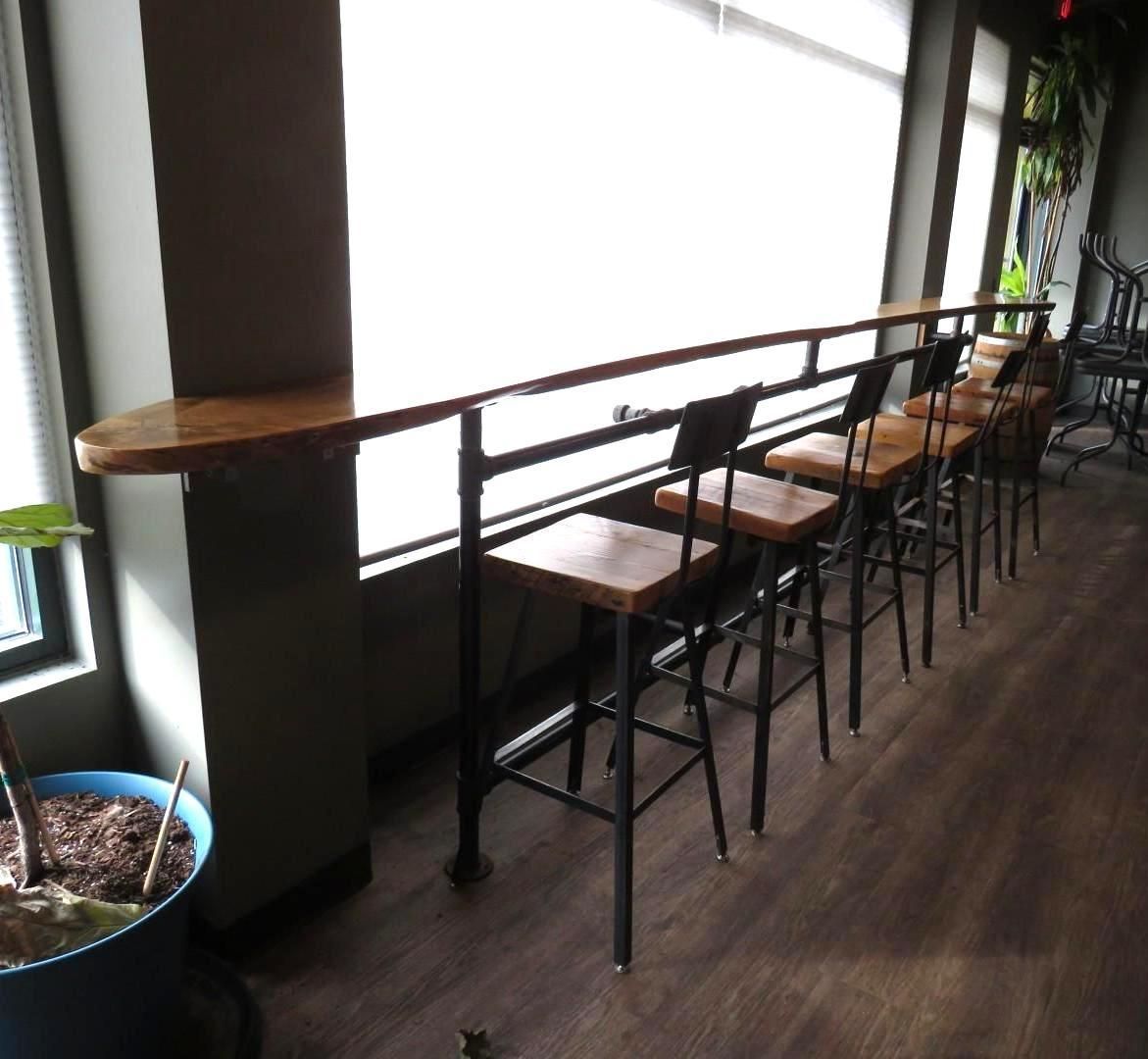 Long wooden bar with black metal stools in front of a large window, natural light.