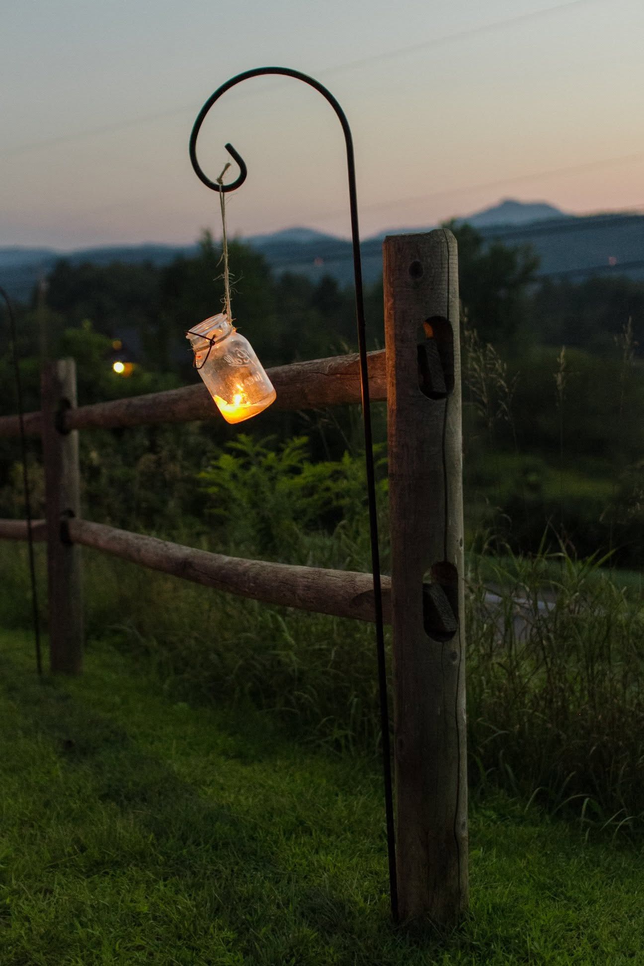Glowing jar lantern hangs from a curved post beside a rustic fence at dusk.