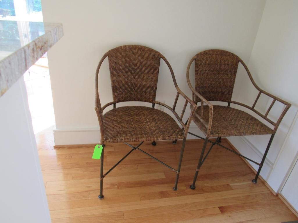 Two brown wicker chairs with metal frames sit on a wooden floor against a white wall.