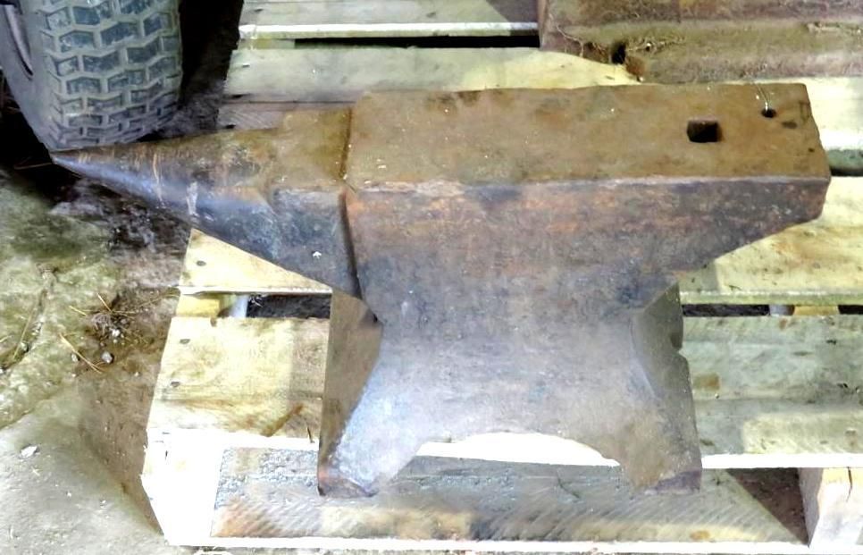 Dark metal anvil on a wooden pallet, with a pointed horn, rectangular work surface, and square and round holes.