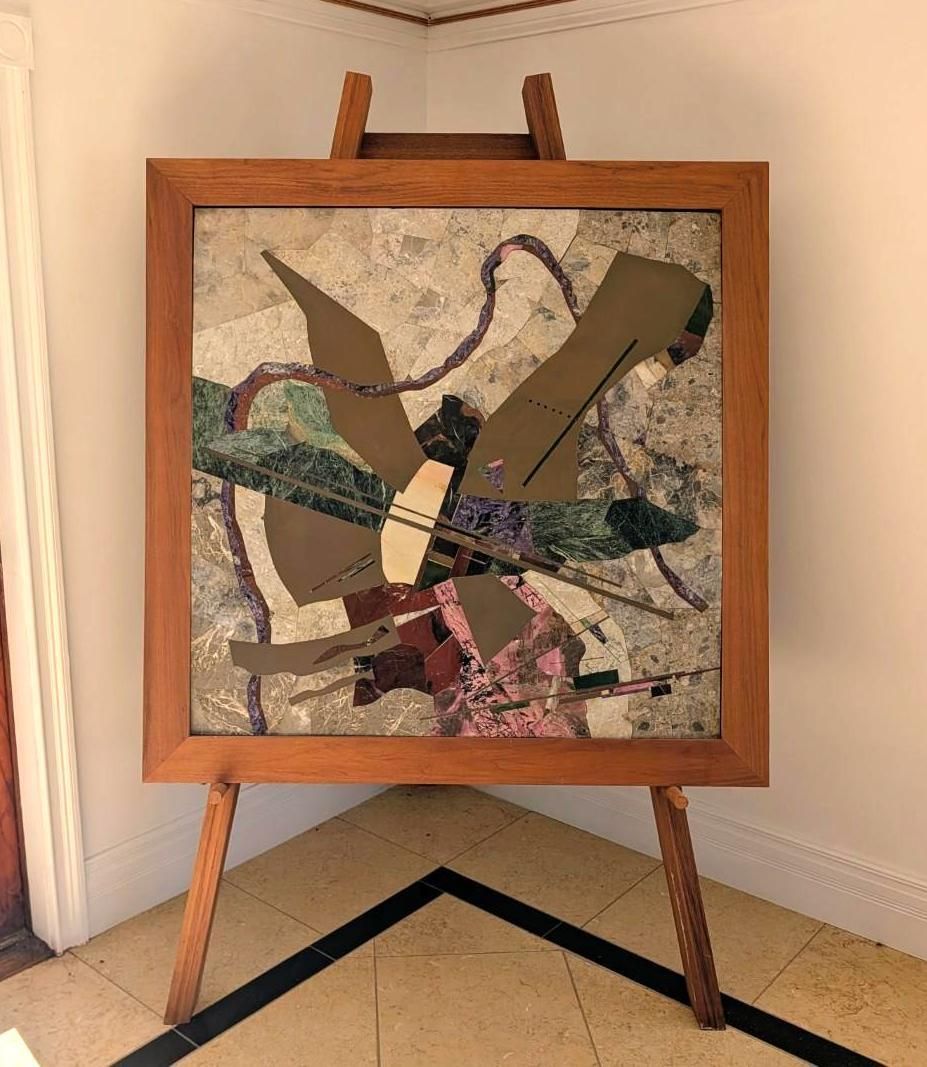 Abstract collage artwork in a wooden easel frame against a beige wall.