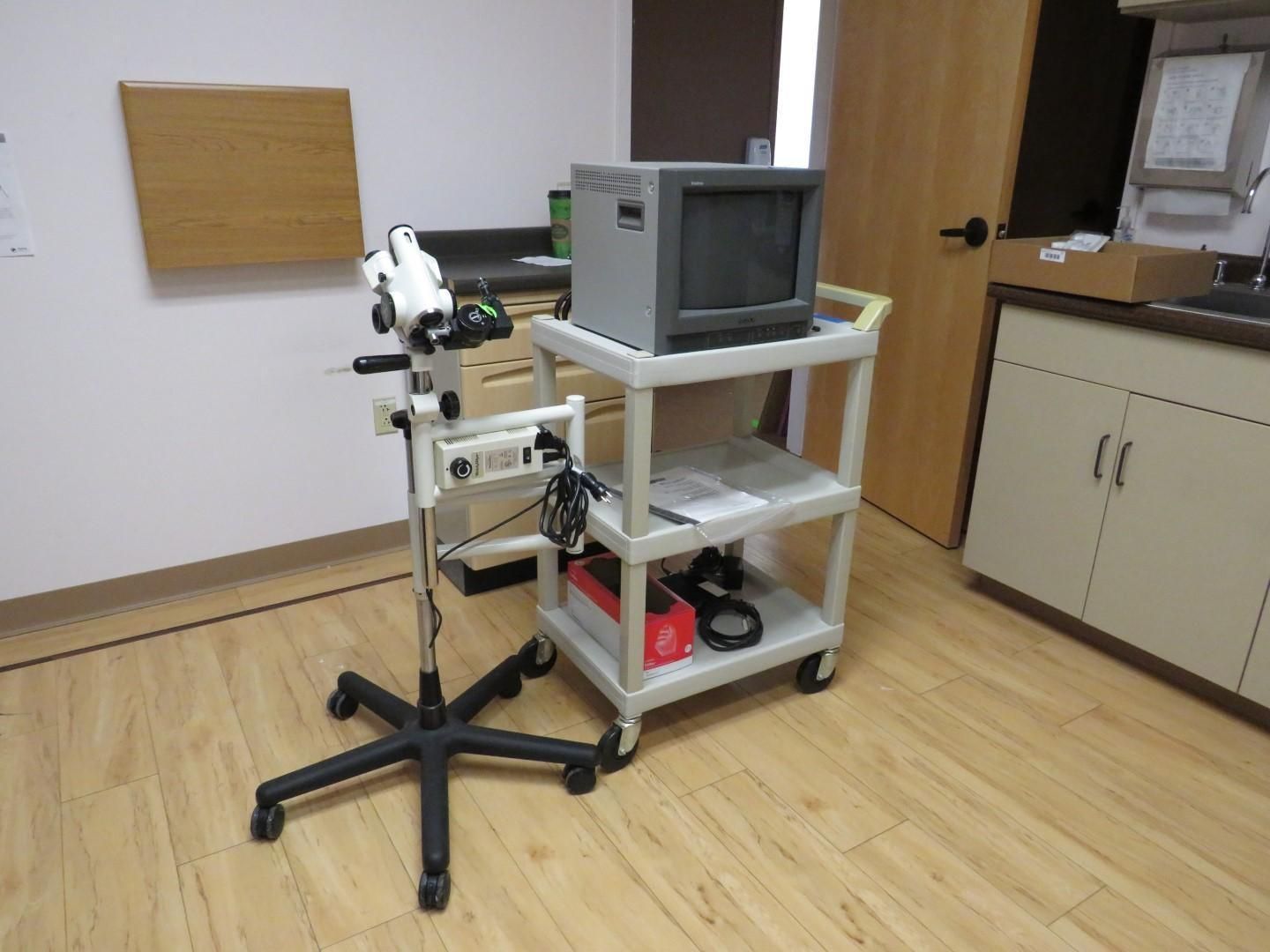 Medical exam room with equipment: microscope, monitor on rolling cart, cabinet, door, and light wood flooring.
