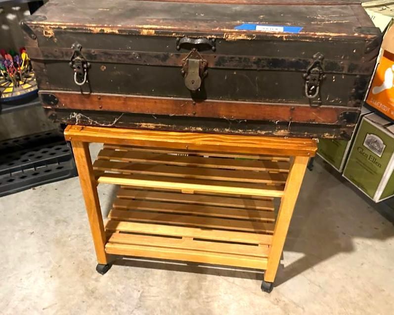 Antique trunk on a wooden cart with shelves, set on a concrete floor.