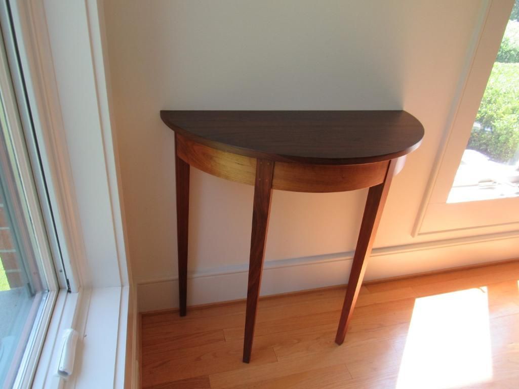 Half-circle table with a dark top and light wood legs, positioned against a white wall near a window.