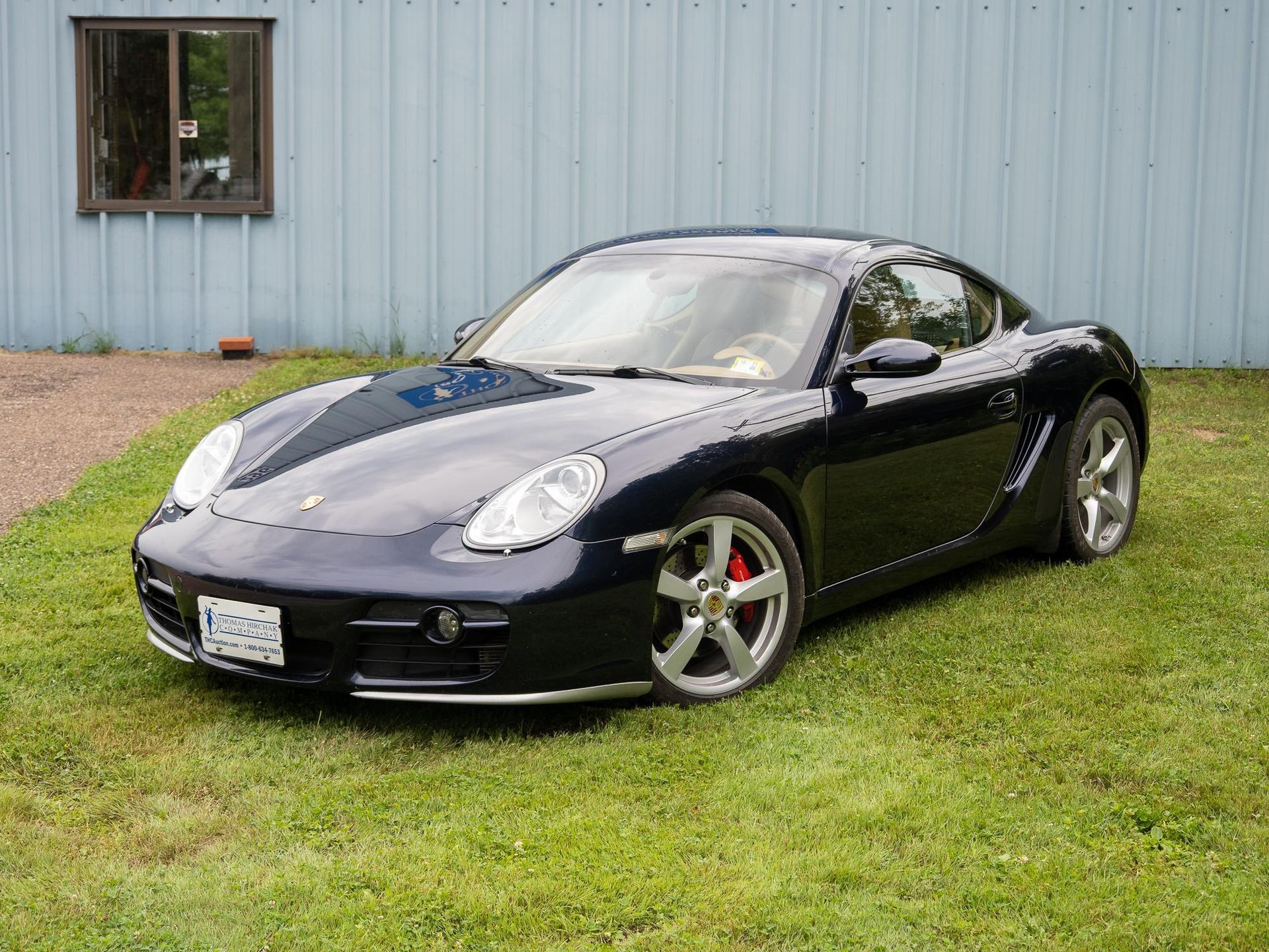 Dark blue Porsche coupe parked on grass in front of a blue building with a small window.