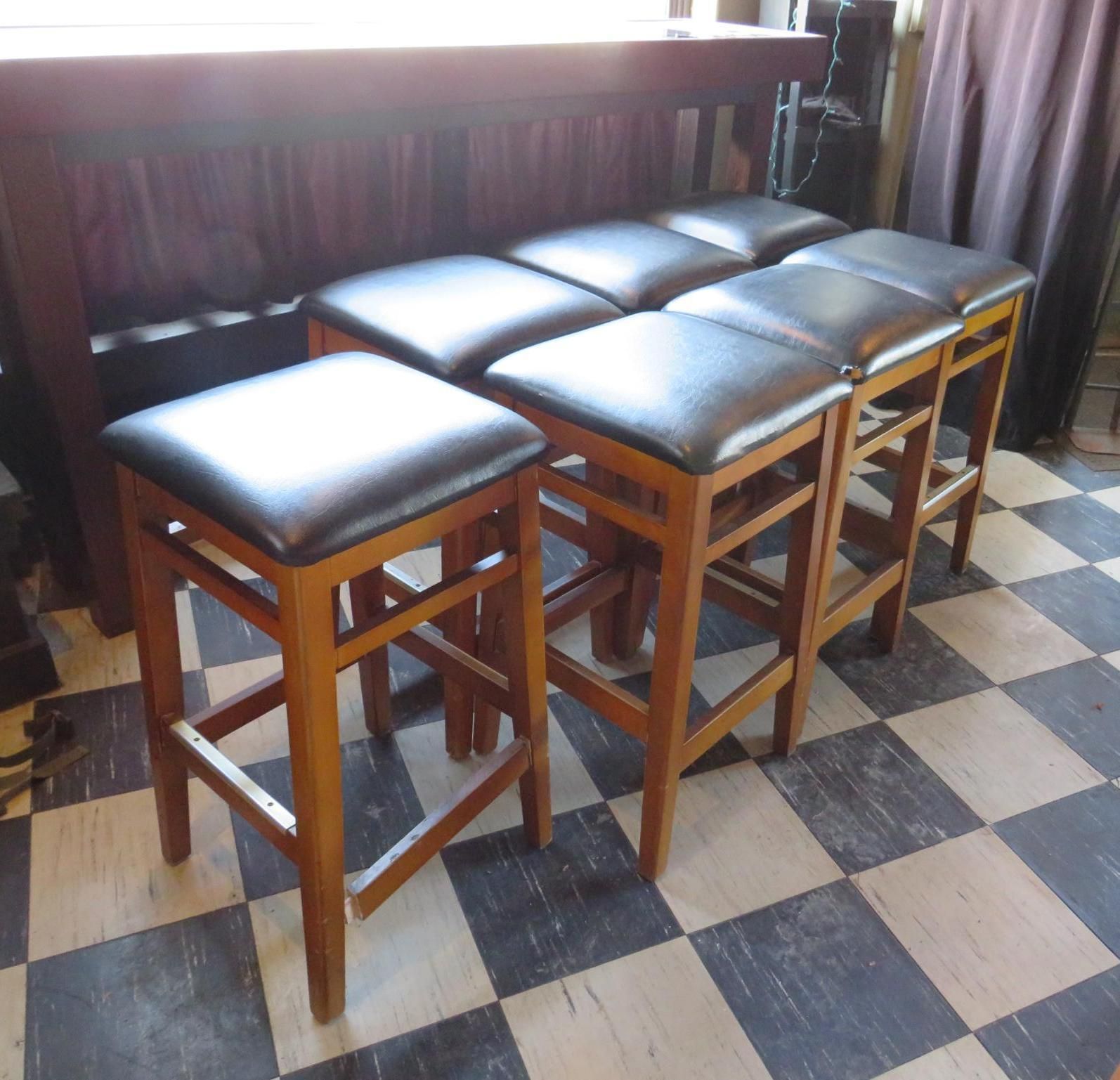 Seven brown wooden bar stools with black cushioned seats arranged in front of a dark wood bar, on a checkered floor.
