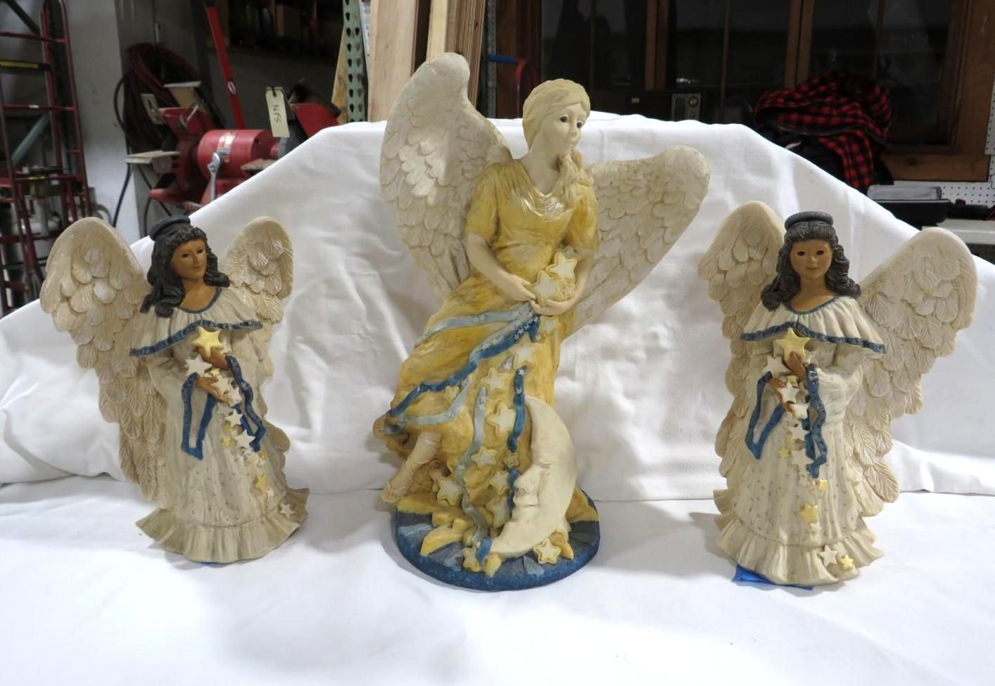Three angel figurines on a white surface; center angel is taller, wearing yellow; side angels hold stars.