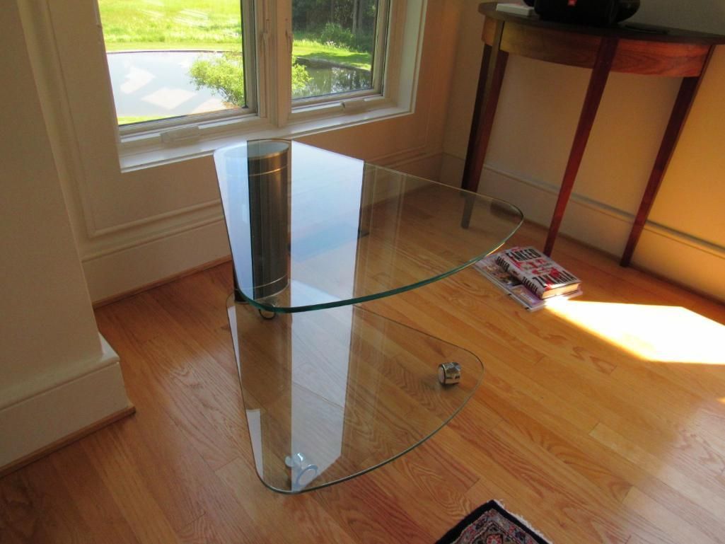 Glass coffee table on wood floor near a window and side table; sunny interior.