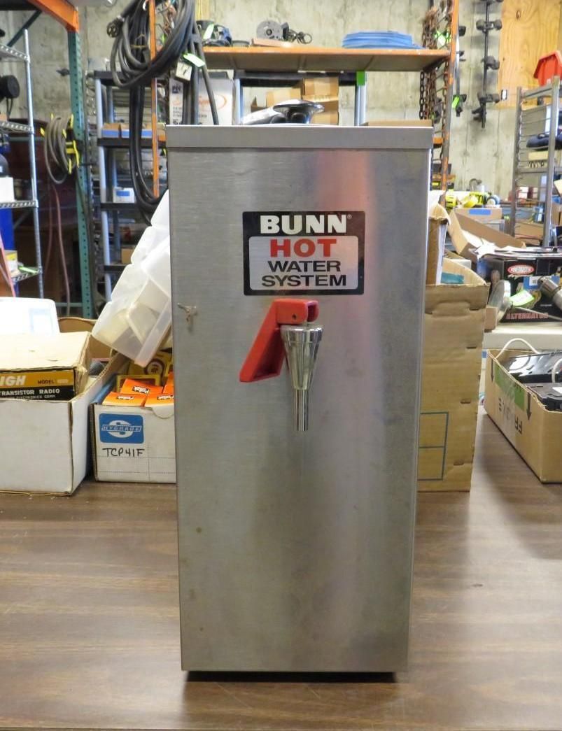 Stainless steel BUNN hot water system with red lever and dispenser, in a storage room.
