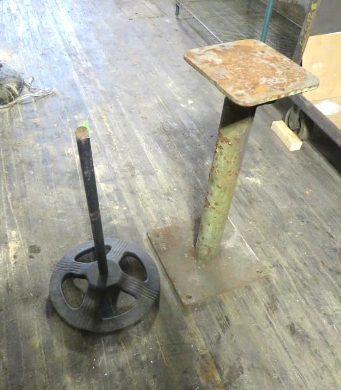 Two metal stands on a wooden floor: a round black stand and a green, rectangular-based stand.