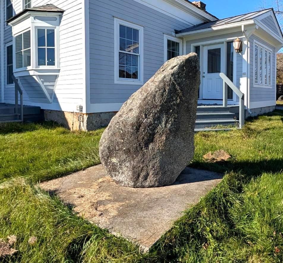 Large upright stone on a concrete pad in a grassy yard beside a light blue house.