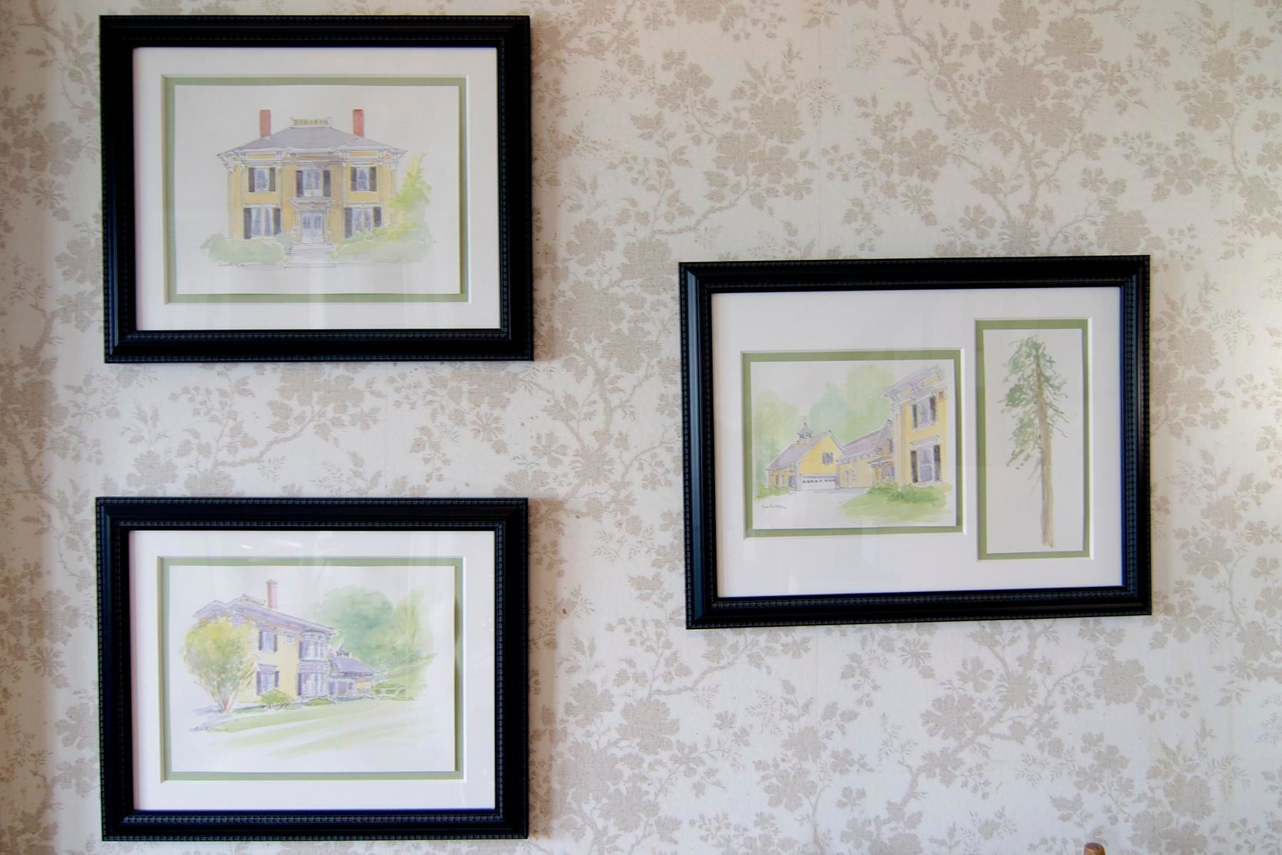 Three framed house drawings on a floral wallpapered wall. Black frames, pastel colors.